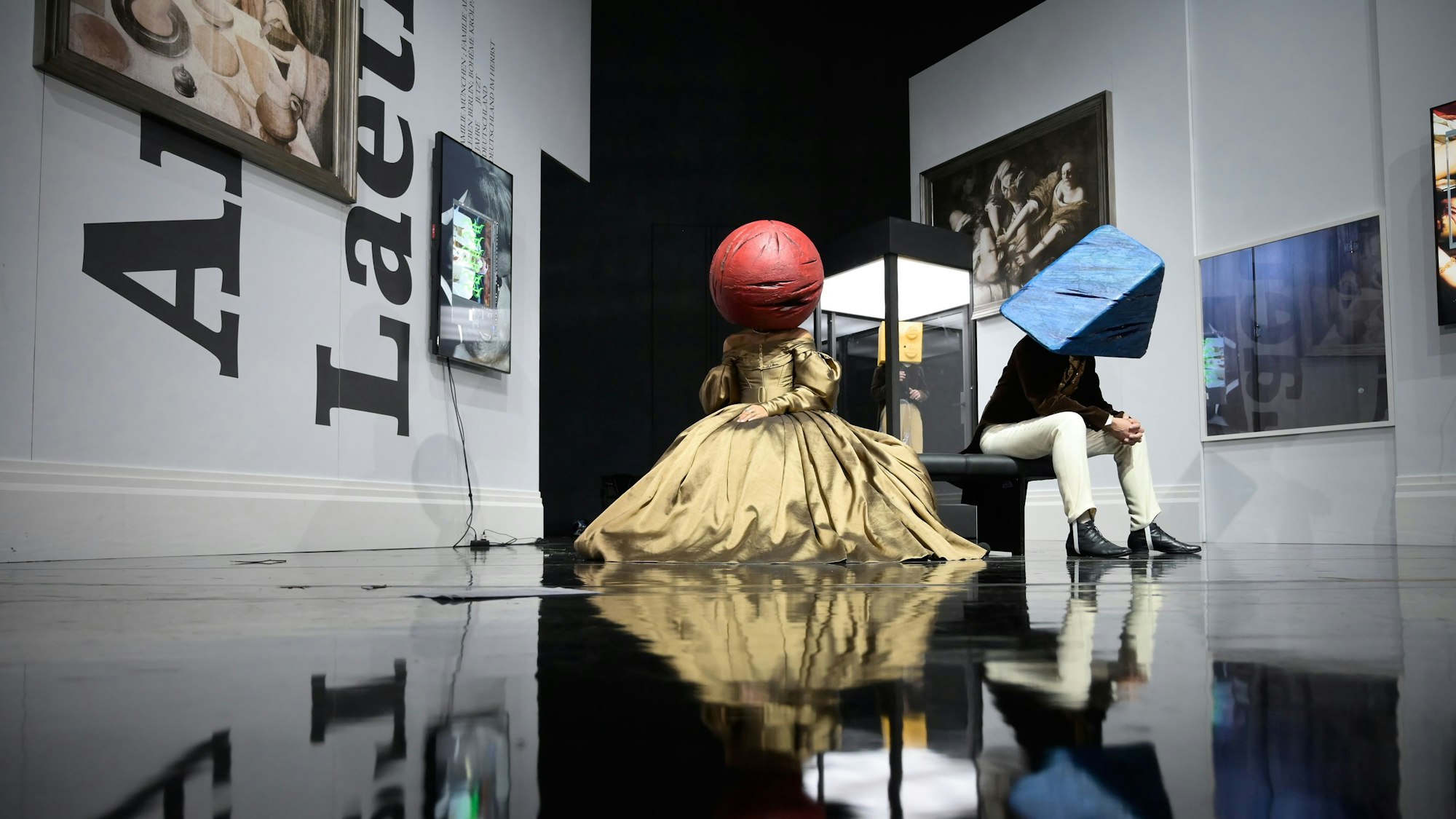 Frau und Mann sitzen in Biedermeier-Kostümen auf einer Bank in der Kulisse von Claudia Bossards „Baracke“-Inszenierung am Deutschen Theater Berlin. Hinten steht zwischen ihnen ihr Kind in einem Glaskasten. Die Frau trägt einen roten Ball über ihren Kopf, der Mann ein blaues Dreieck, das Kind einen gelben Legostein-Quader.