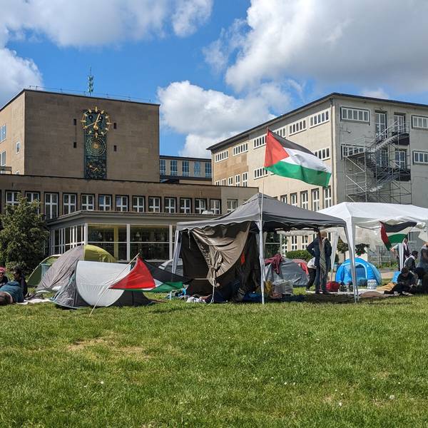 Zelte und ein Pavillon mit Palästina-Flaggen stehen auf der Uniwiese, im Hintergrund ist das Hauptgebäude der Uni Köln zu sehen.