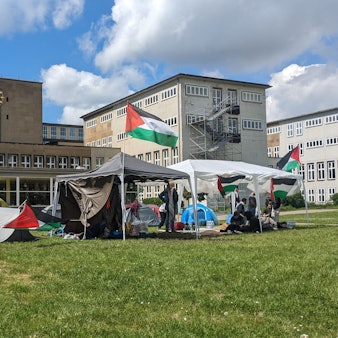 Zelte und ein Pavillon mit Palästina-Flaggen stehen auf der Uniwiese, im Hintergrund ist das Hauptgebäude der Uni Köln zu sehen.