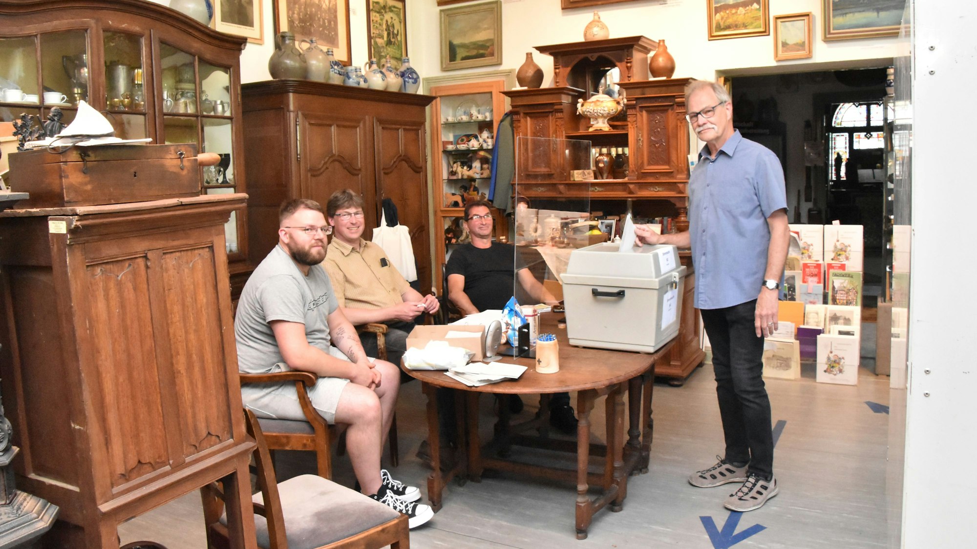 Drei Männer sitzen hinter einer Wahlurne in einem historisch eingerichteten Zimmer. Ein Mann wirft seinen Stimmzettel in die Urne.