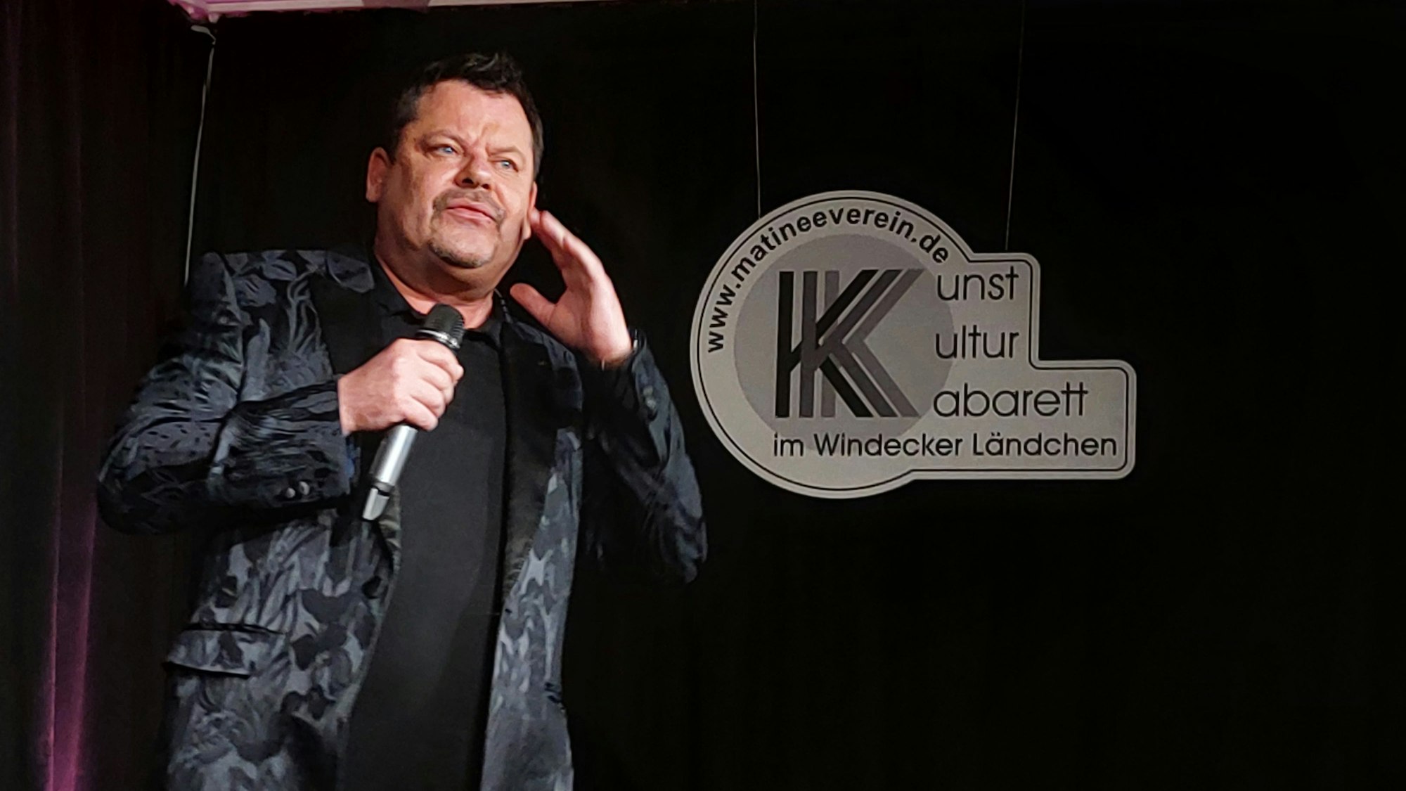 Ein Mann im gemusterten grau-schwarzen Sakko; er hat ein Mikrophon in der Hand. Im Hintergrund hängt ein Schild des Matineevereins. Kunst, Kultur, Kabarett im Windecker Ländchen steht darauf.