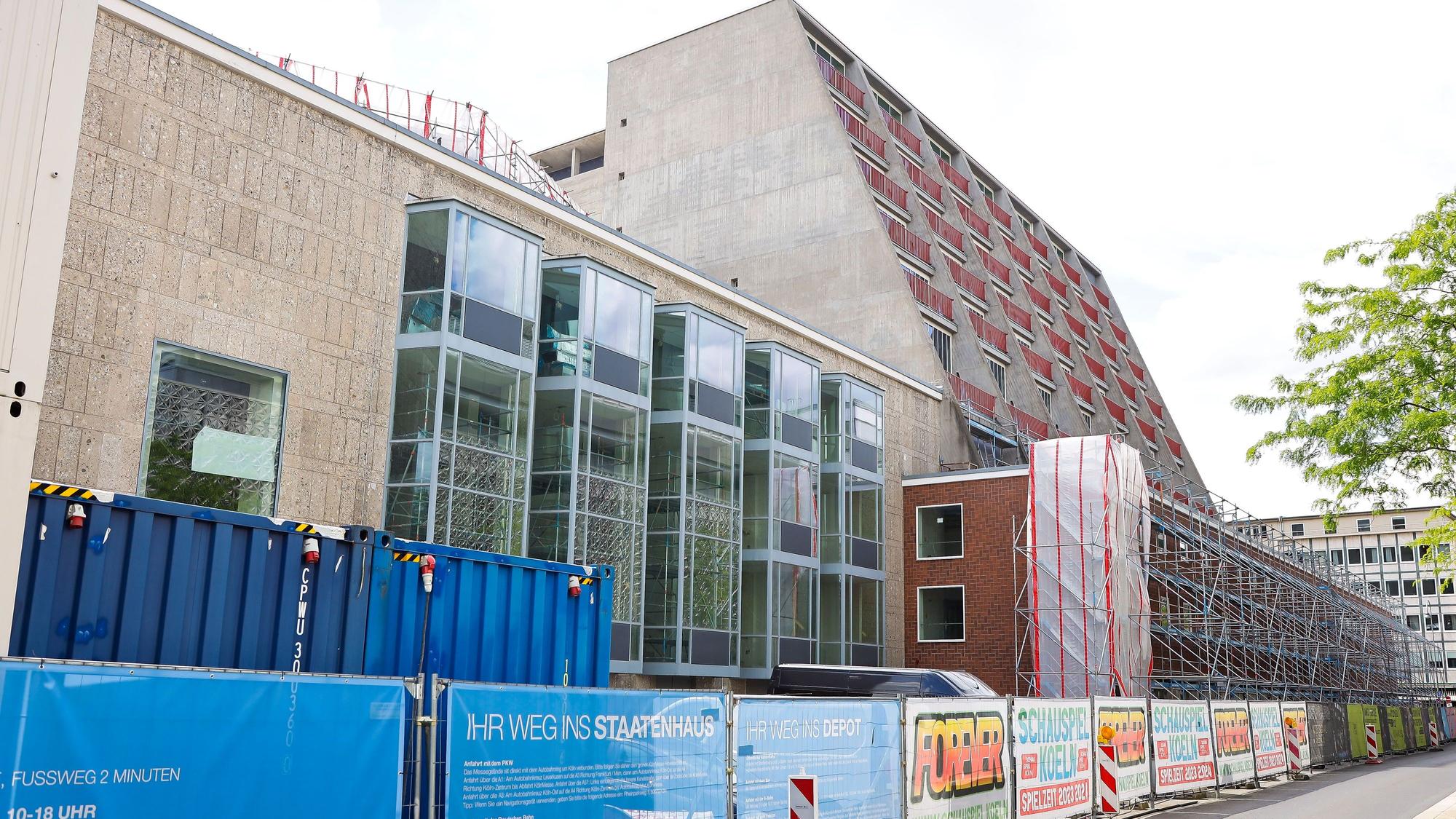 Der Eingang zur Kölner Oper am Offenbachplatz wird von Baugerüsten verdeckt. Bauzäune mit blauen Plastikplanen und Werbung für das Programm der städtischen Bühnen grenzen das Gebäude vom Offenbachplatz ab.