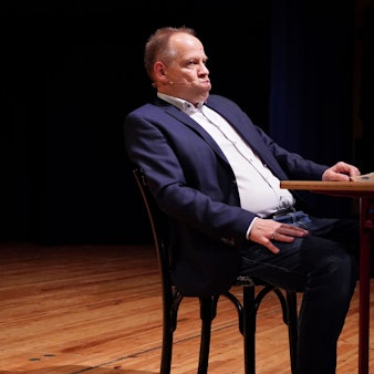 Kabarettist Thomas Schreckenberger sitzt im Stadttheater in Euskirchen an einem Tisch. Er gibt eine gelungene Version von Klaus Kinski zum besten.