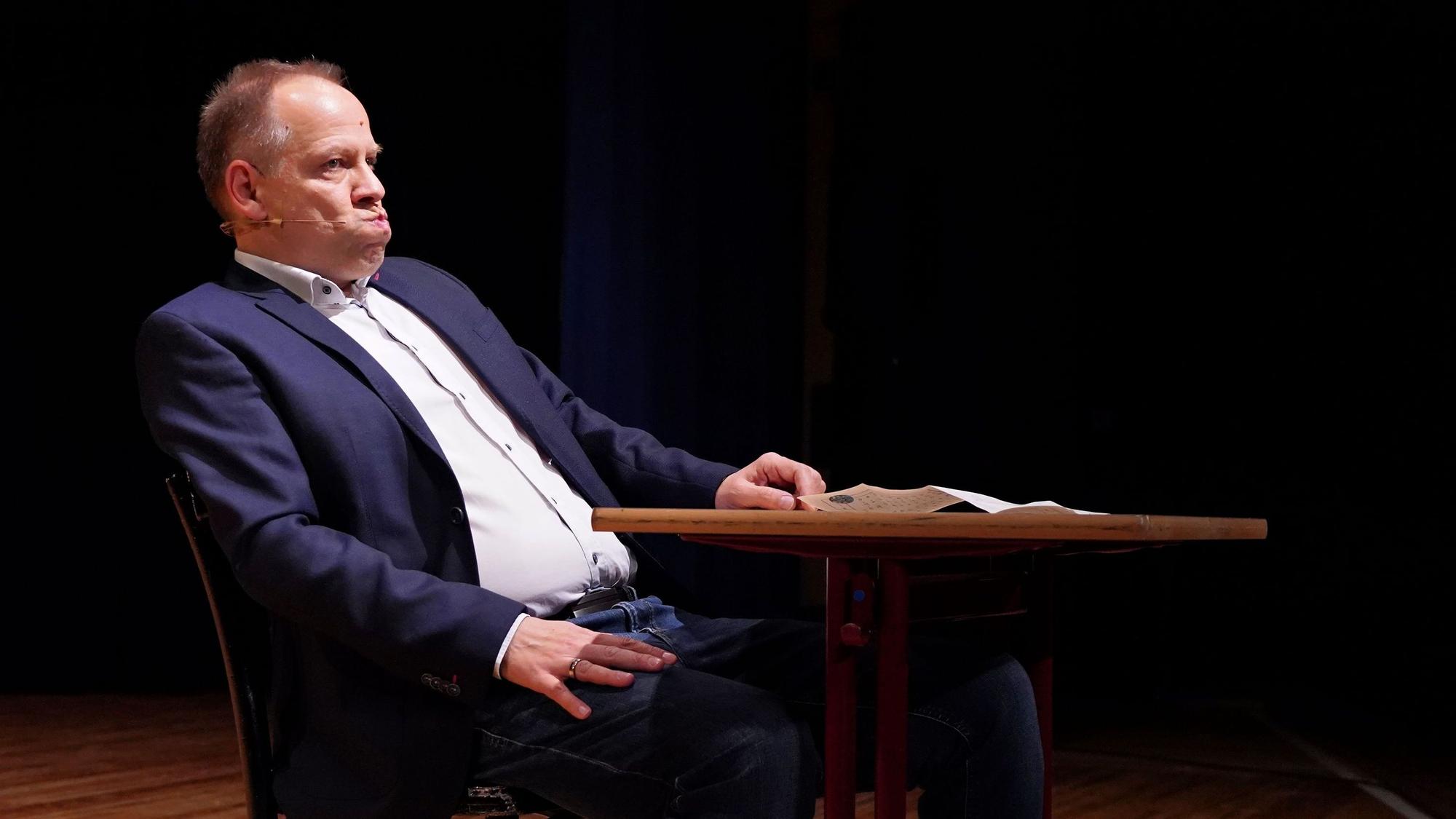 Kabarettist Thomas Schreckenberger sitzt im Stadttheater in Euskirchen an einem Tisch. Er gibt eine gelungene Version von Klaus Kinski zum besten.