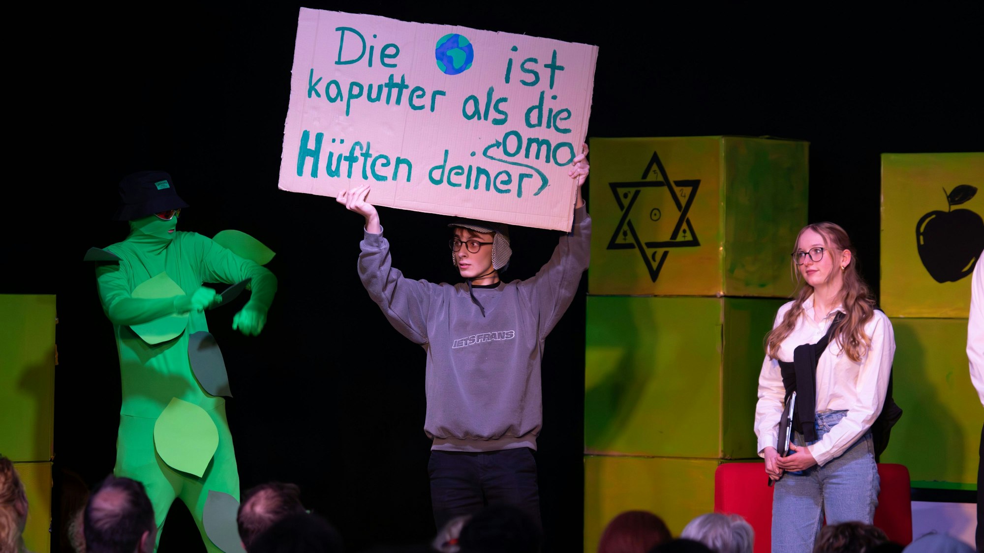 Drei Schüler stehen auf der Bühne des Lessing-Gymnasiums. In der Mitte hält ein Schüler ein Schild hoch, auf dem steht: „Die Welt ist kaputter als die Hüften deiner Oma“. Foto von René Denzer