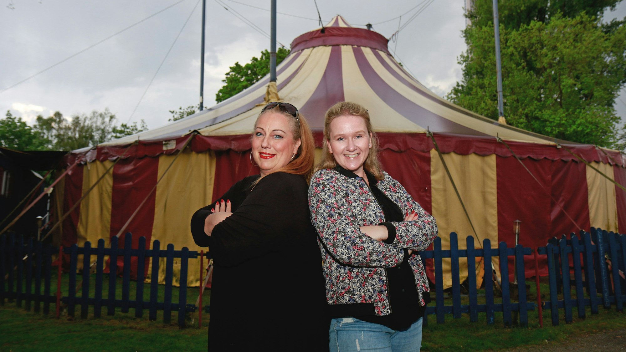 Zwei Frauen lehnen Rücken an Rücken gegeneinander. Im Hintergrund ein Zirkuszelt.