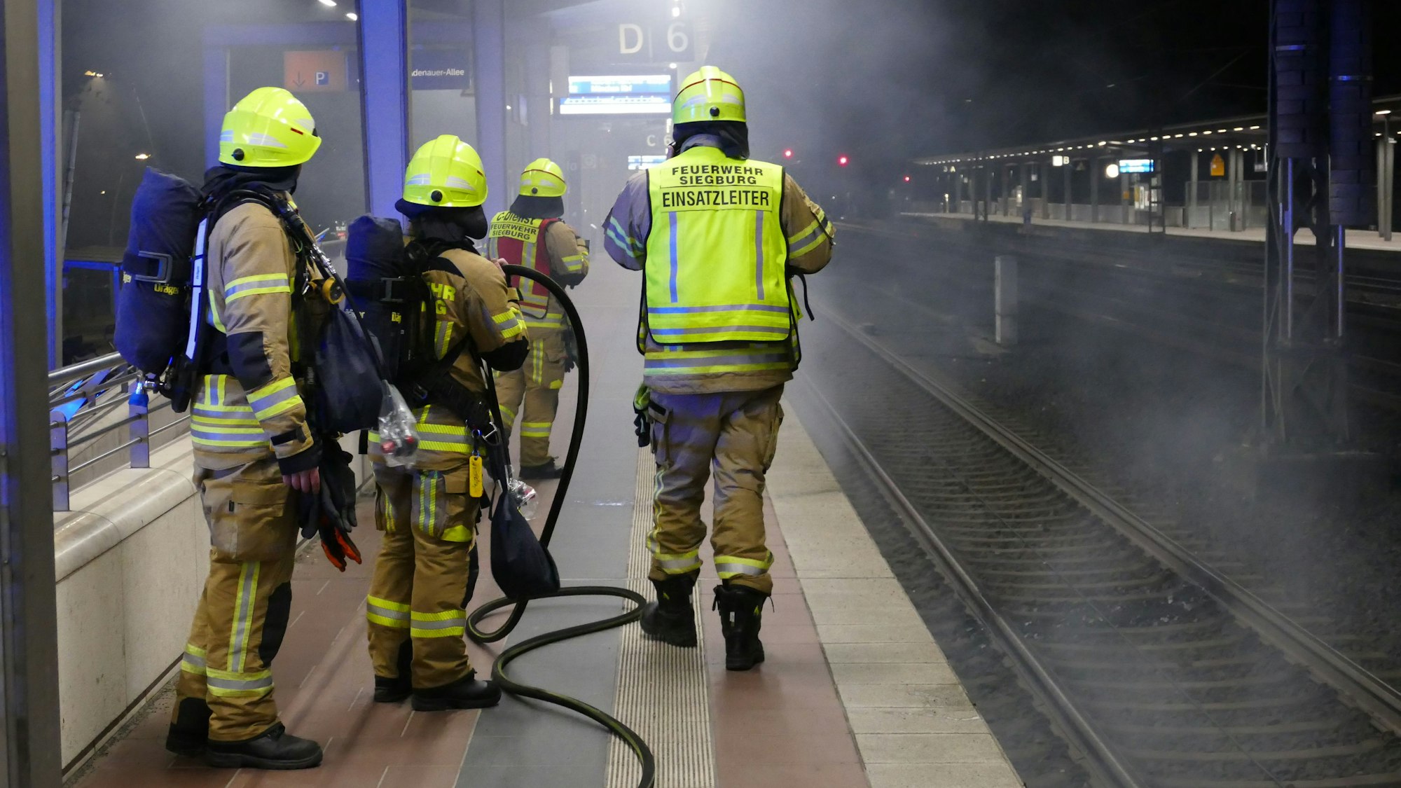 Feuerwehrleute stehen am Bahnsteig, unter dem Rauch hervordringt.