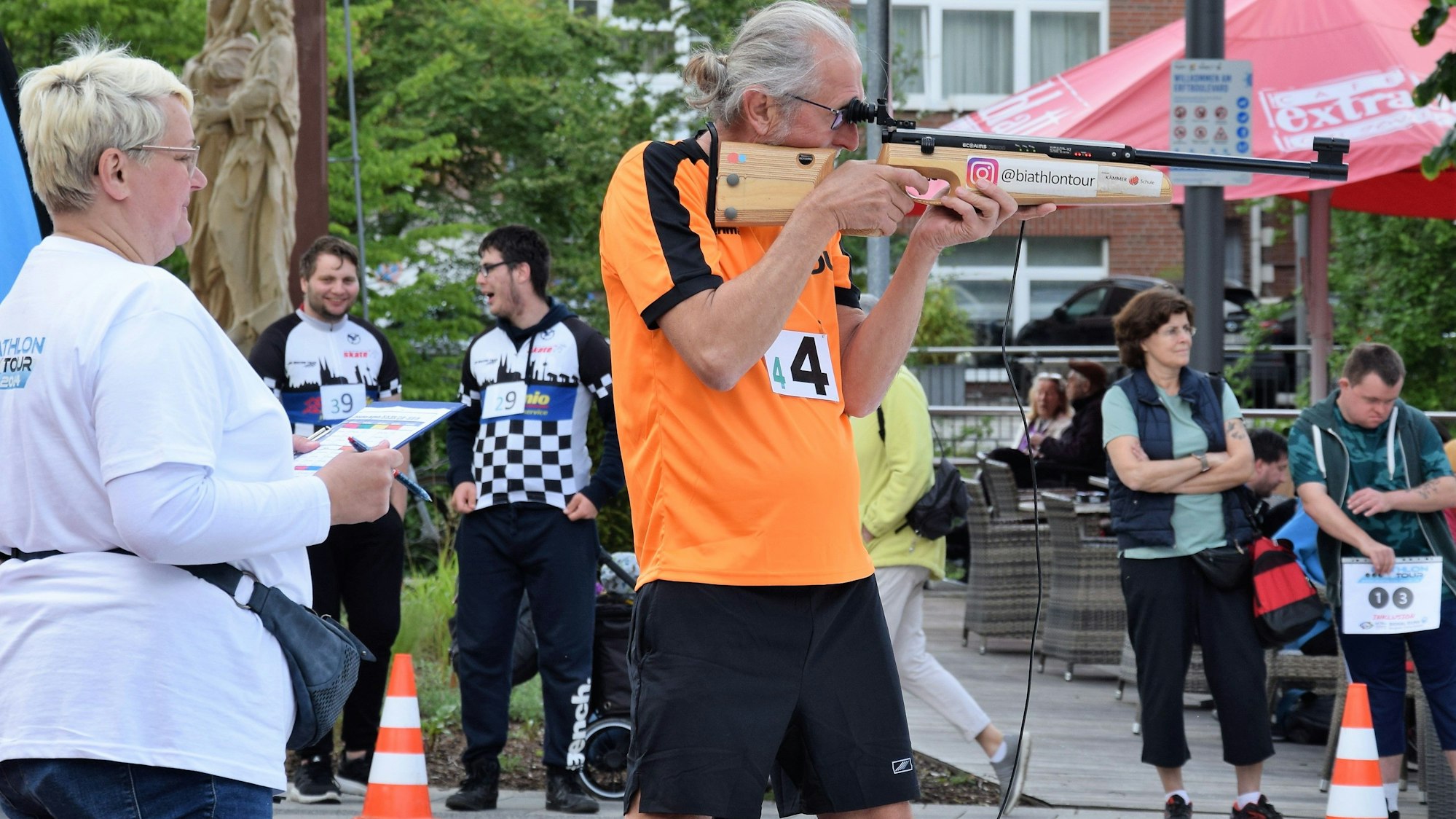 Ein Mann mit grauen Haaren schießt mit einem Gewehr an einem Schießstand, mehrere Menschen schauen zu.