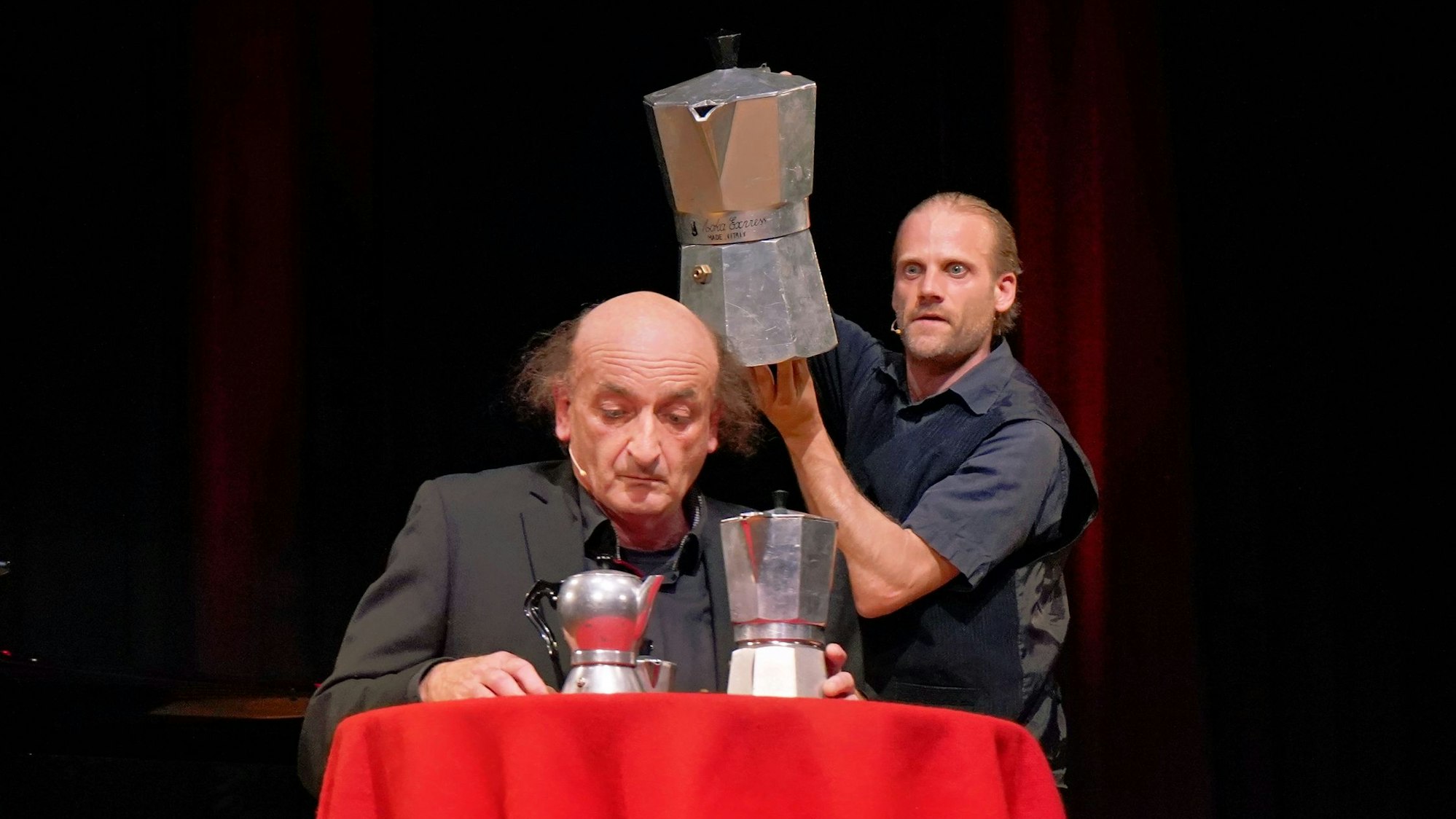 Das Duo Rado und Schaller tritt im Stadttheater Euskirchen auf. Einer sitzt an einem mit einer roten Decke überzogenen Tisch, der andere hält eine überdimensionale Kaffeekanne über seinen Kopf.