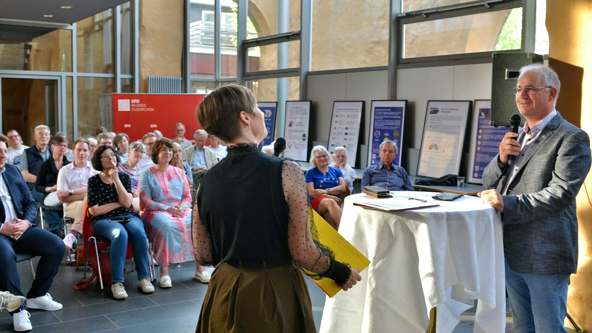 Annegret Lewak und Helmut Dahmen, beide mit Mikrofon, im Gespräch, im Hintergrund das Publikum.