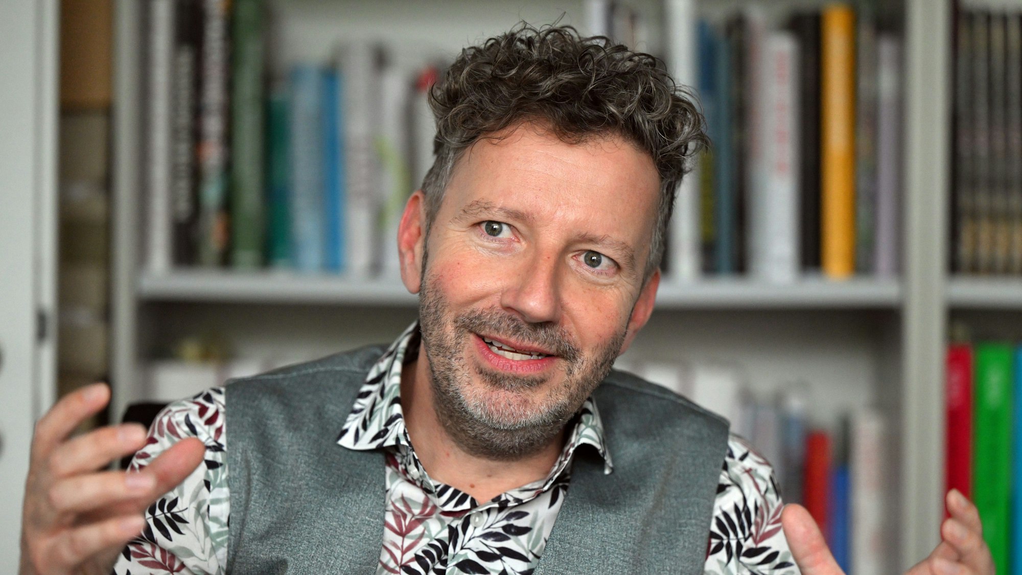 Thorsten Schorn sitzt vor einem Bücherregal. Er trägt ein Hemd mit Blumenmuster und eine graue Weste