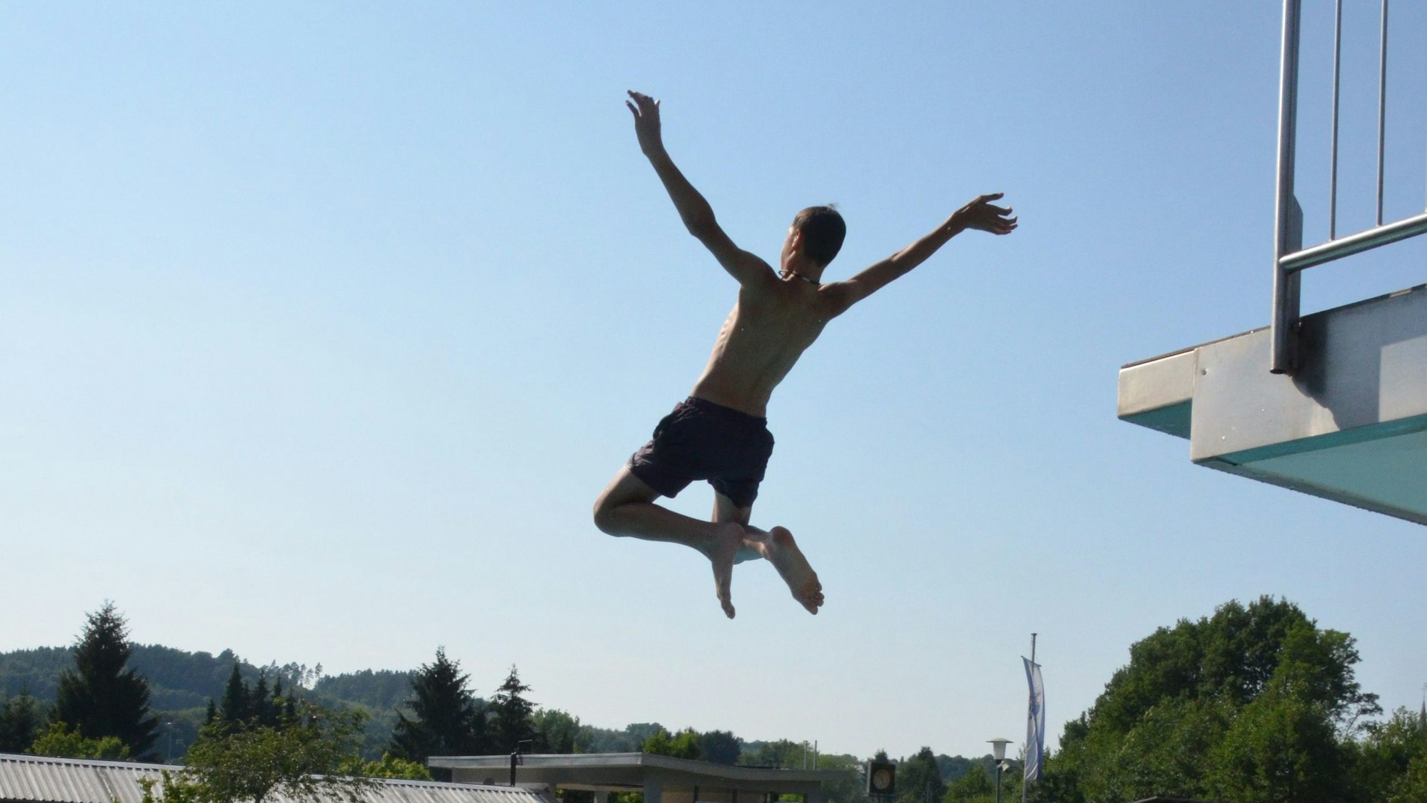 Eine Person springt in ein Schwimmbecken. Im Wasser befinden sich Besucher. Im Hintergrund Häuser und Bäume.