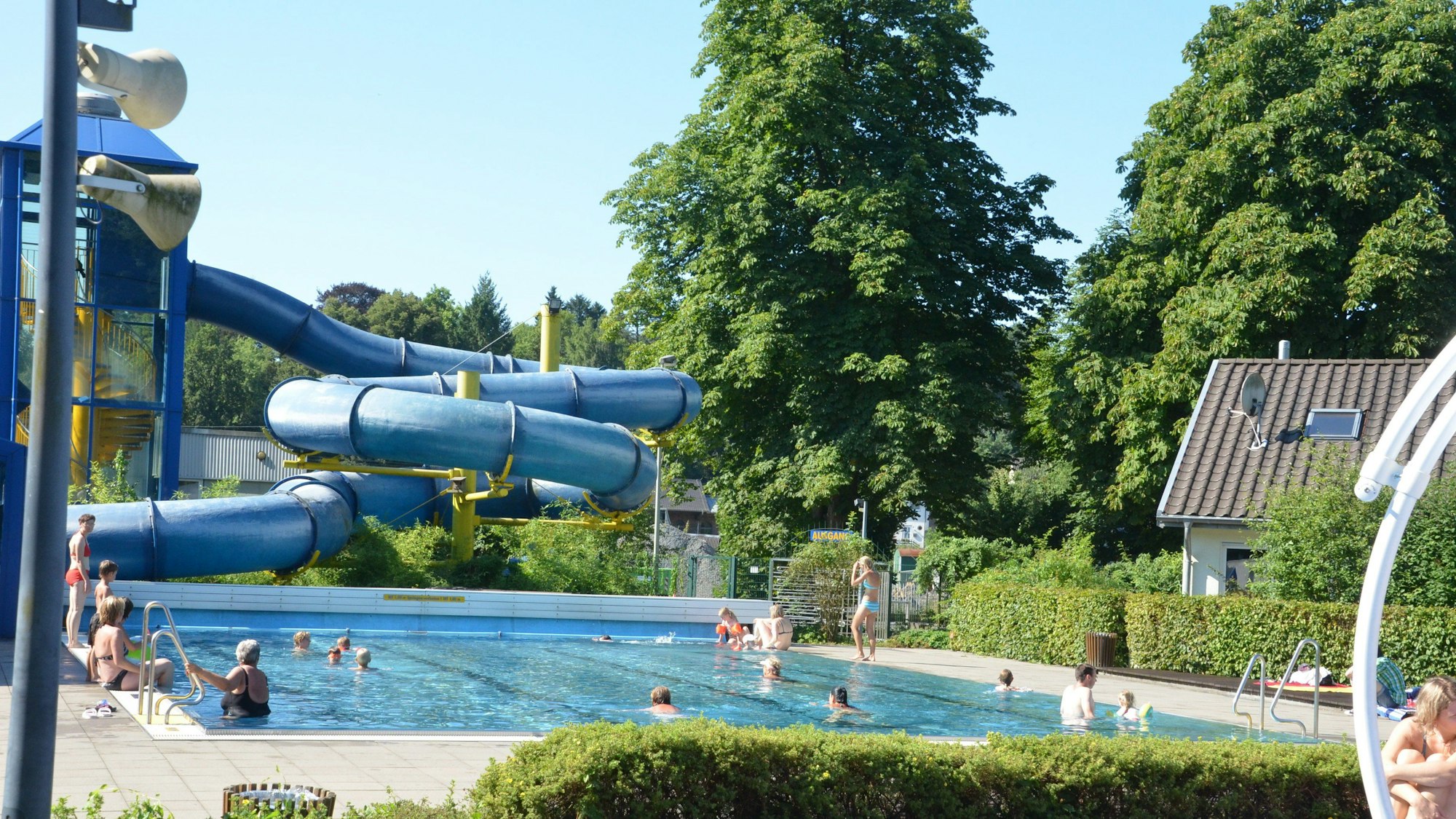Ein Schwimmbecken mit Besuchern. Im Hintergrund eine blaue Rutsche und Bäume.