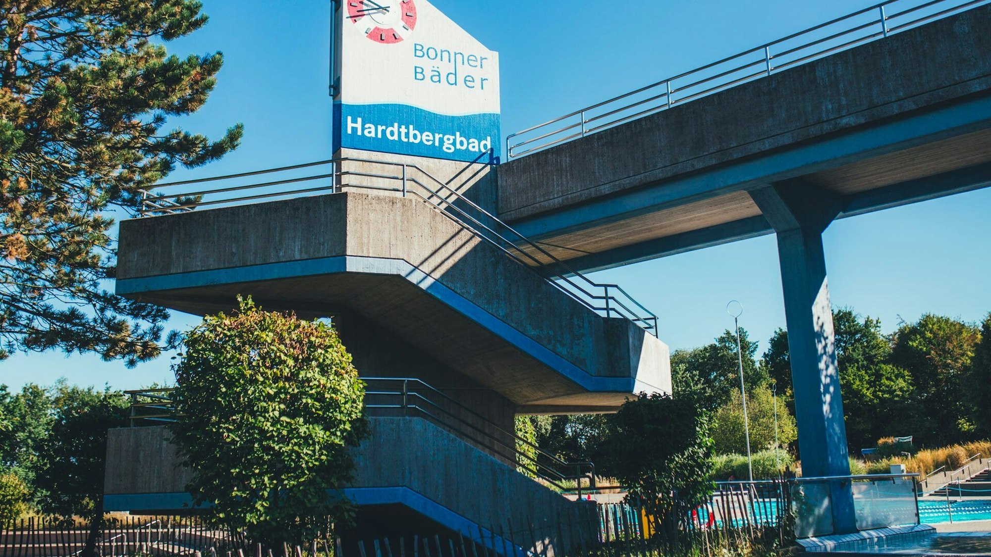 Eine Brücke mit dem Schild und der Aufschrift „Hardtbergbad“ ist zu sehen. Im Hintergrund sieht man ein Schwimmbecken.