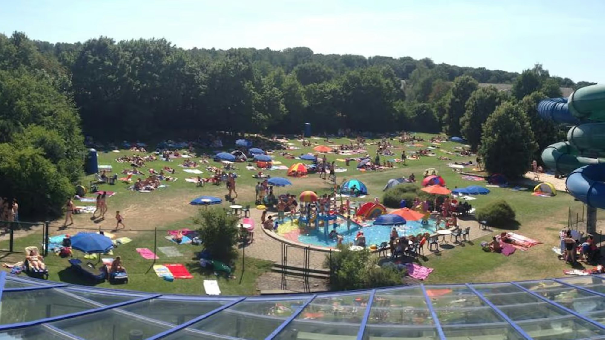 Ein Blick von einem Vordach aus auf eine gut besuchte Freibadfläche mit Bäumen im Hintergrund.