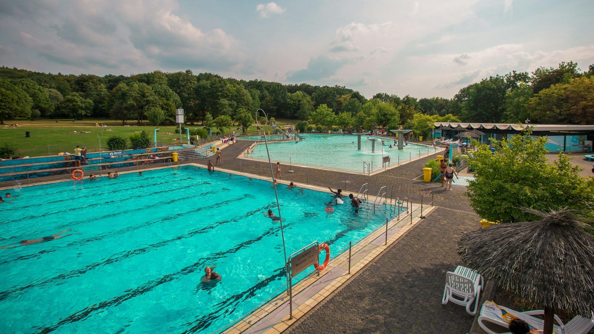 Ein großes Schwimmbecken mit acht Schwimmbahnen und ein kleineres Schwimmbecken liegen nebeneinander. Im Hintergrund sind eine Wiese und Bäume.