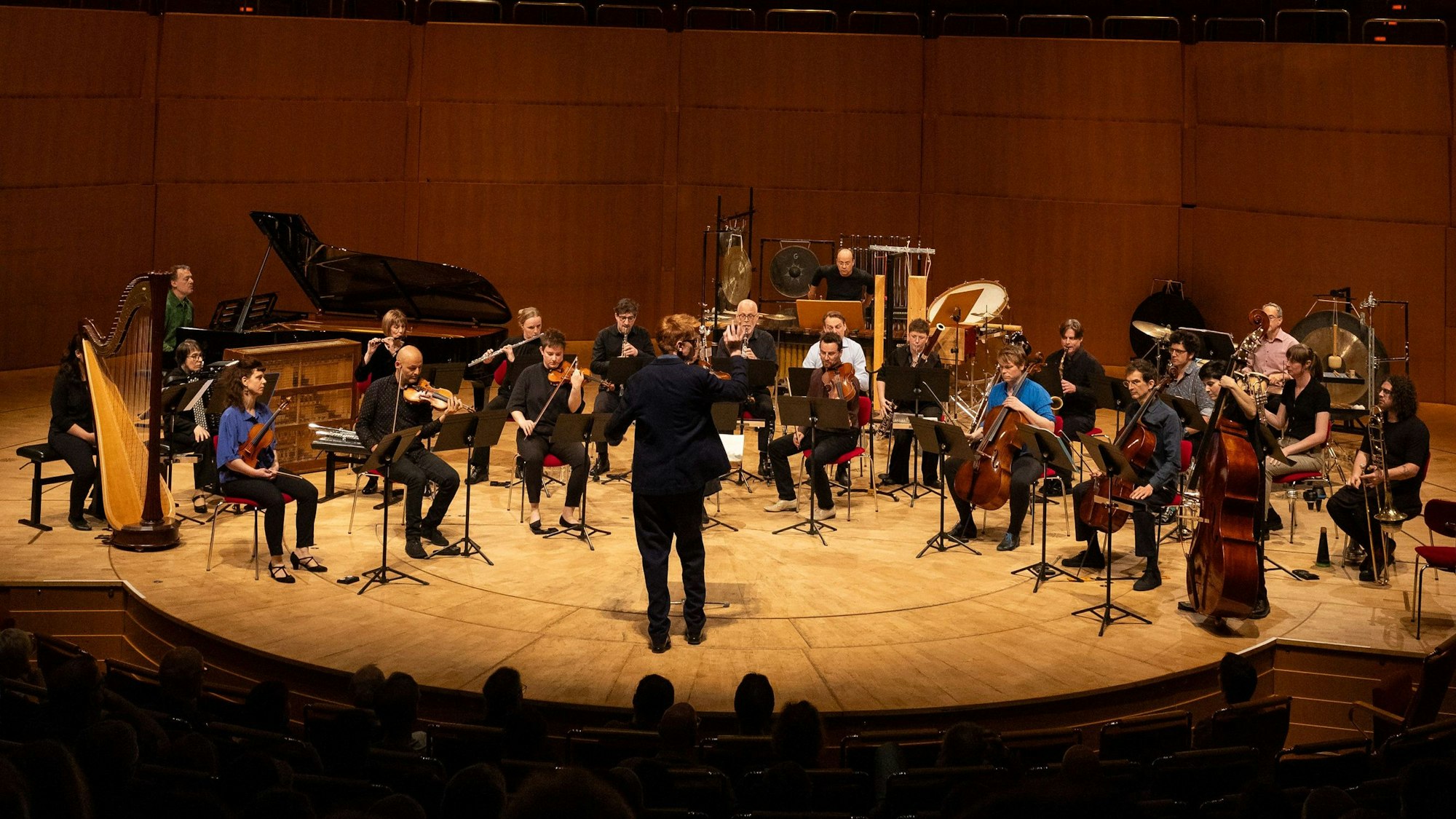 Auf der Bühne der Philharmonie steht das Ensemble mit einem Flügel, einer Harfe, Perkussion, Streichern und Bläsern. Vorne steht der Dirigent Enno Poppe, dem Ensemble zugewandt.