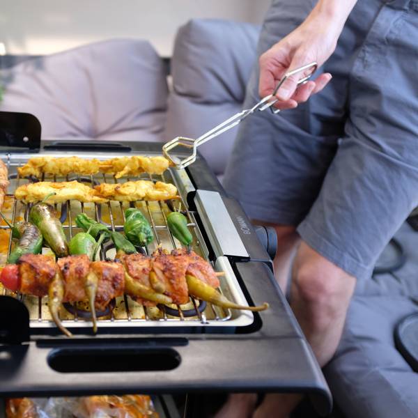 Ein Mann, der eine Grillzange hält, brät auf seinem Balkon mithilfe eines Elektrogrills Fleisch und Gemüse