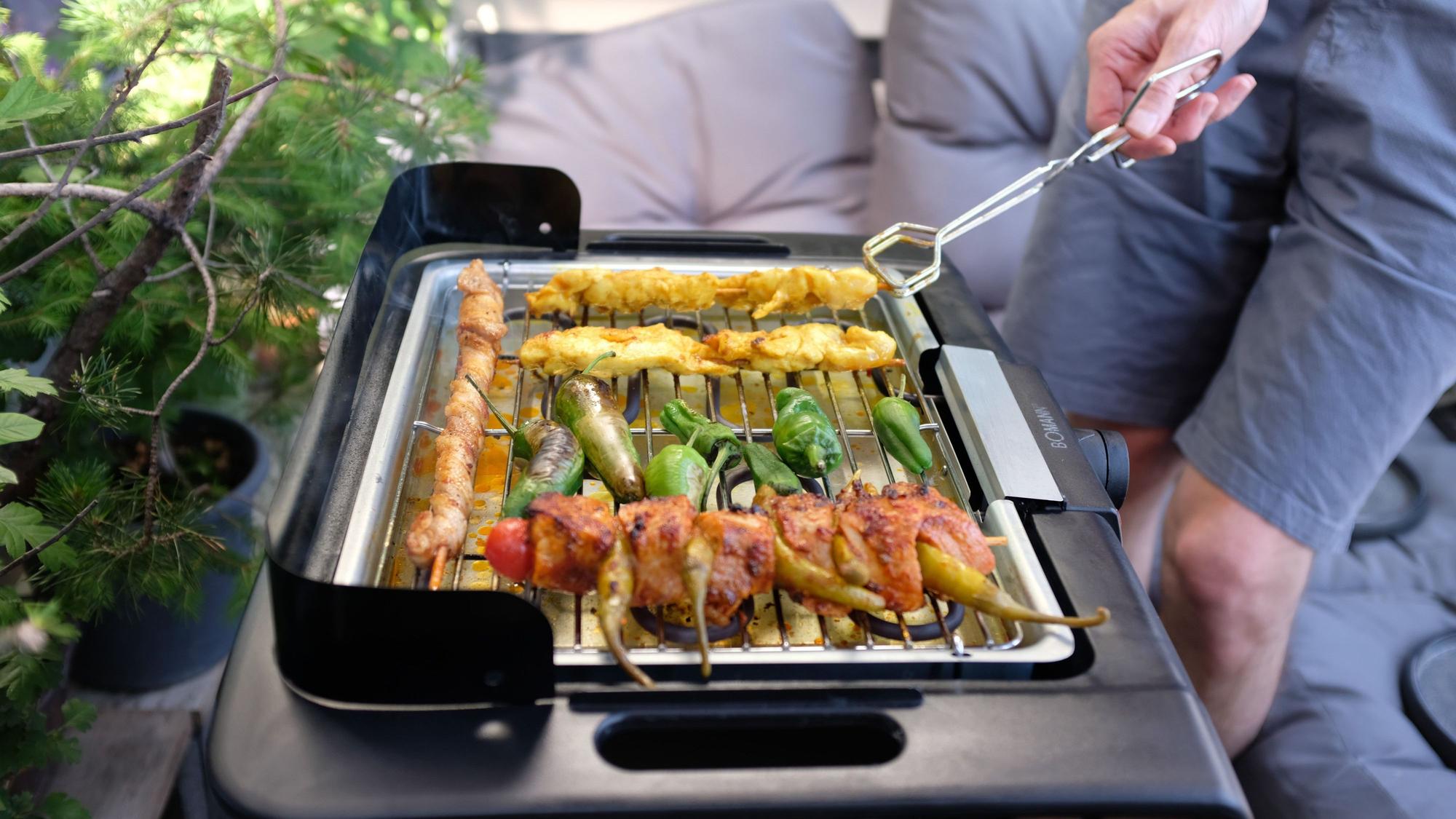Ein Mann, der eine Grillzange hält, brät auf seinem Balkon mithilfe eines Elektrogrills Fleisch und Gemüse