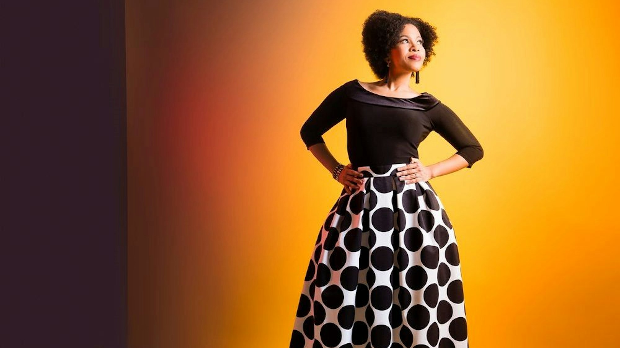 Die aus Südafrika stammende Star-Sopranistin Golda Schultz steht vor einer gelben Wand und trägt einen Rock mit großen schwarzen Punkten, am 19. Mai feiert sie ihr Debüt in Köln als Solistin bei einem Benefizkonzert des Gürzenich-Orchesters in der Philharmonie.