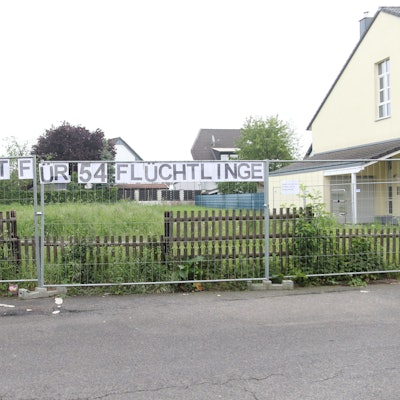Über einem Zaun hängt ein Transparent mit der Aufschrift „Ein guter Ort für 54 Flüchtlinge“ und daneben ein Zettel mit der Aufschrift „Sagen uns die Stadt Pulheim und der Bürgermeister“.