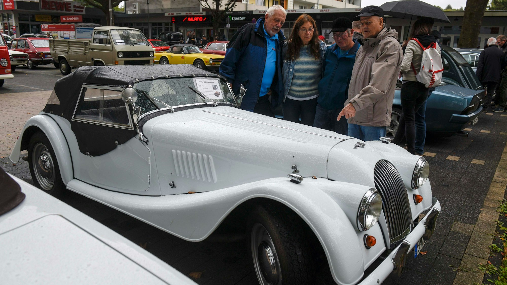 Zu sehen ist ein alter Wagen mit geschwungenen Kotflügeln, der von Menschen bestaunt wird.