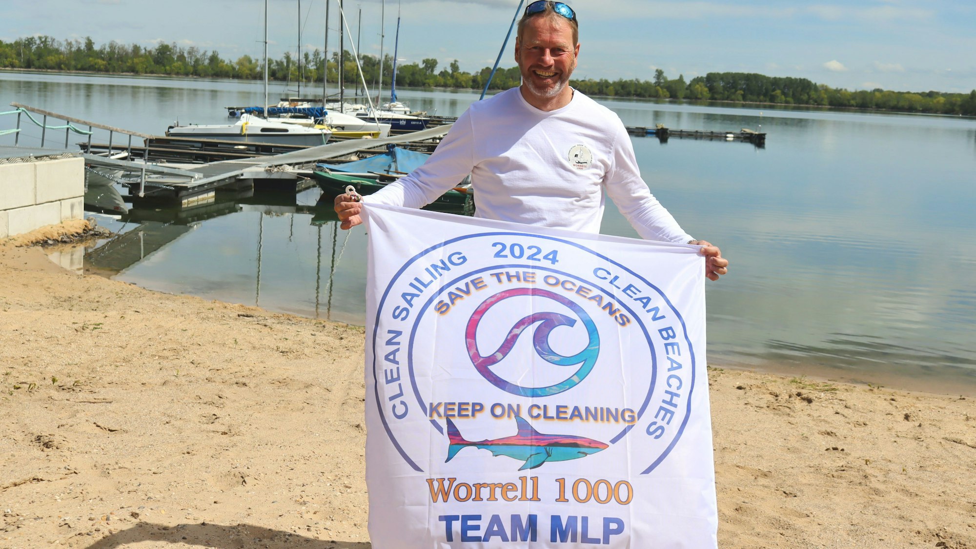 André Hauschke hält eine Fahne hoch, auf der „Clean Sailing 2024 Clean Beaches, Save the Oceans, Keep on Cleaning“ und der Hinweis „Worrell 1000 - Team MLP“ stehen. Im Hintergrund sind auf einem See Segelboote zu erkennen.