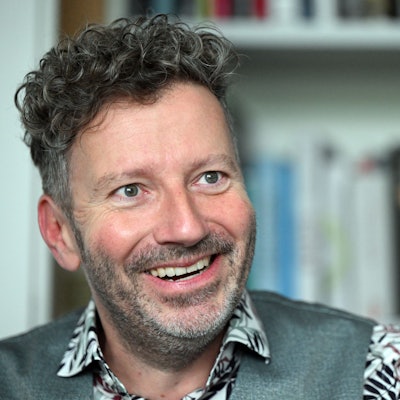 Thorsten Schorn sitzt vor einem Bücherregal. Er trägt ein Hemd mit Blumenmuster und eine graue Weste