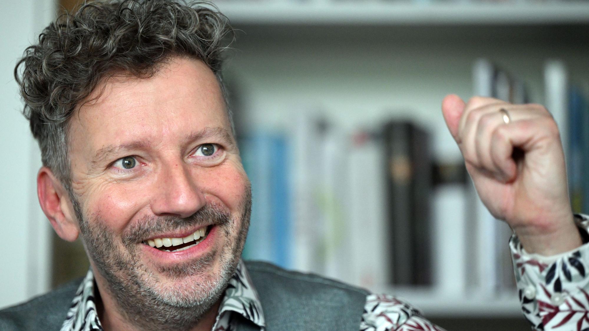 Thorsten Schorn sitzt vor einem Bücherregal. Er trägt ein Hemd mit Blumenmuster und eine graue Weste
