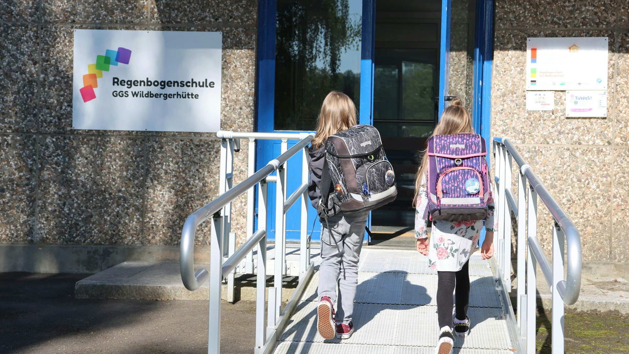 Zwei Mädchen mit Schulranzen sind von hinten zu sehen, wie sie auf die Eingangstüre einer Schule zugehen.