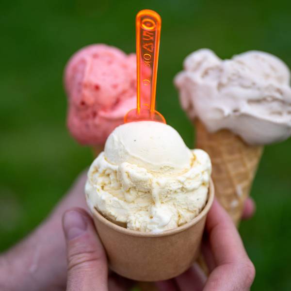 Eine Person hält drei Eis in der Hand, zwei in der Waffel, eine im Becher mit einem orangefarbenen Löffel.