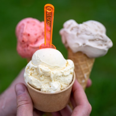 Eine Person hält drei Eis in der Hand, zwei in der Waffel, eine im Becher mit einem orangefarbenen Löffel.