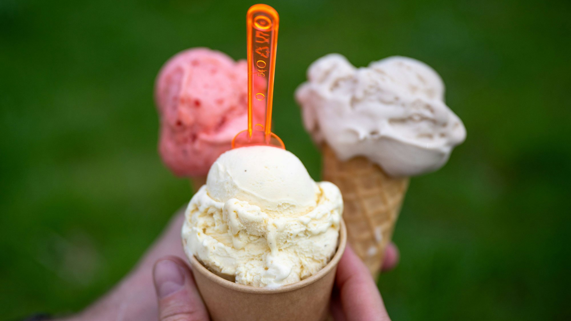 Eine Person hält drei Eis in der Hand, zwei in der Waffel, eine im Becher mit einem orangefarbenen Löffel.