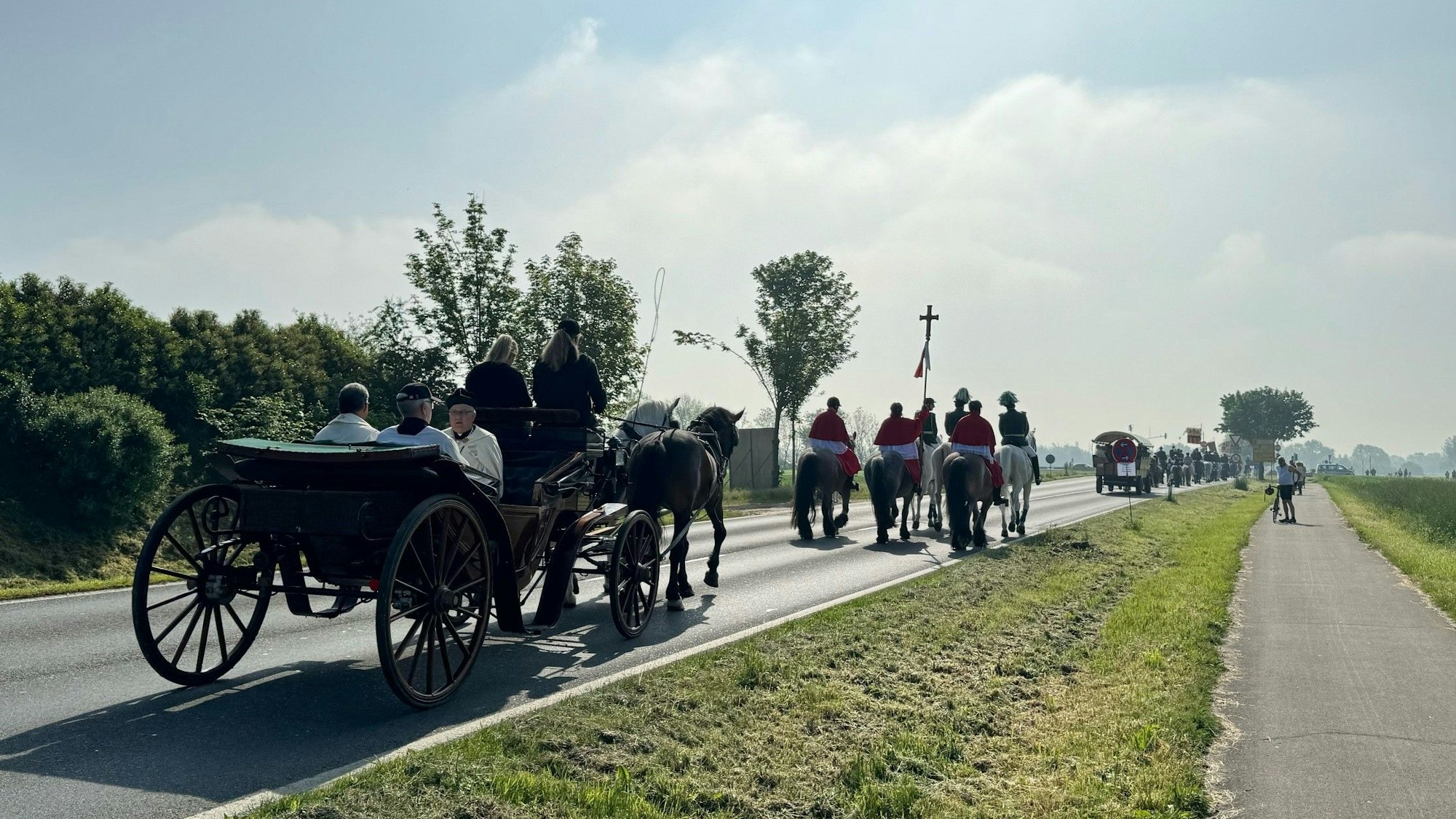 Zu sehen sind Reiter und Kutschen, die auf einer Straße unterwegs sind.