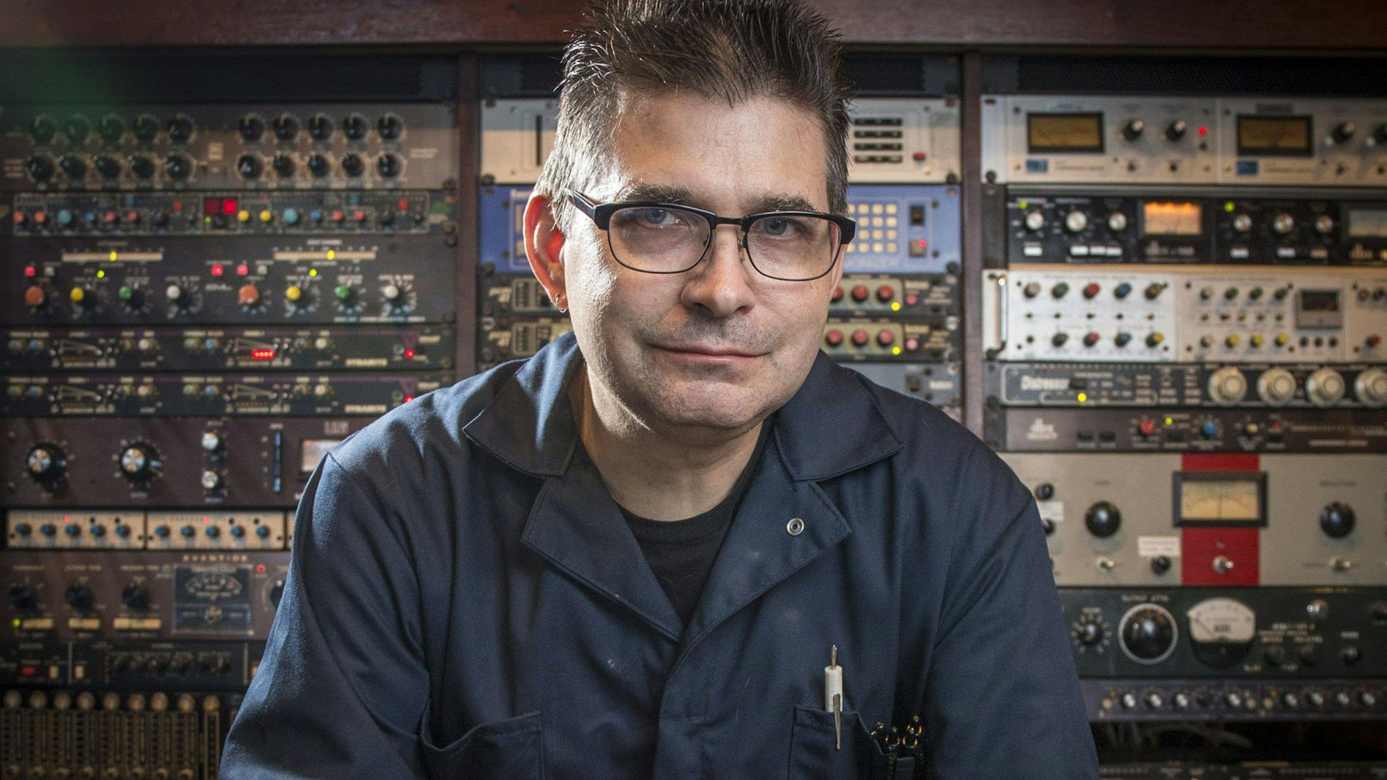 Musikproduzent Steve Albini sitzt im groben blauen Arbeitshemd vor einer Wand aus Geräten in seinem Studio in Chicago.