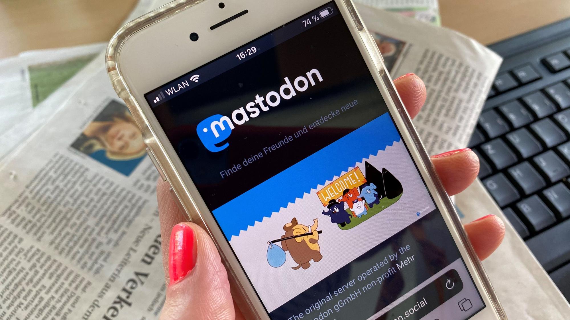 Eine Frau hält ein Smartphone in der Hand, auf dem Bildschirm ist die Startseite des sozialen Netzwerks Mastodon zu erkennen.