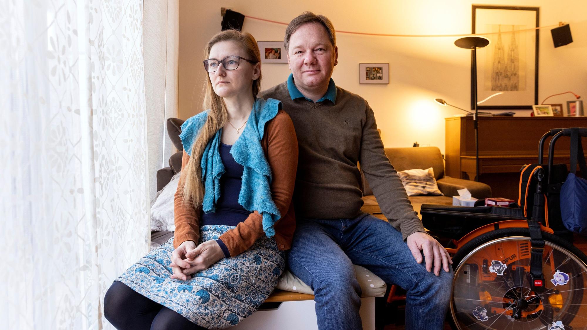Tanja Bock-Schweizer und Sebastian Bock sitzen auf der Sofakante in ihrem Wohnzimmer. Neben ihnen steht ein Rollstuhl.