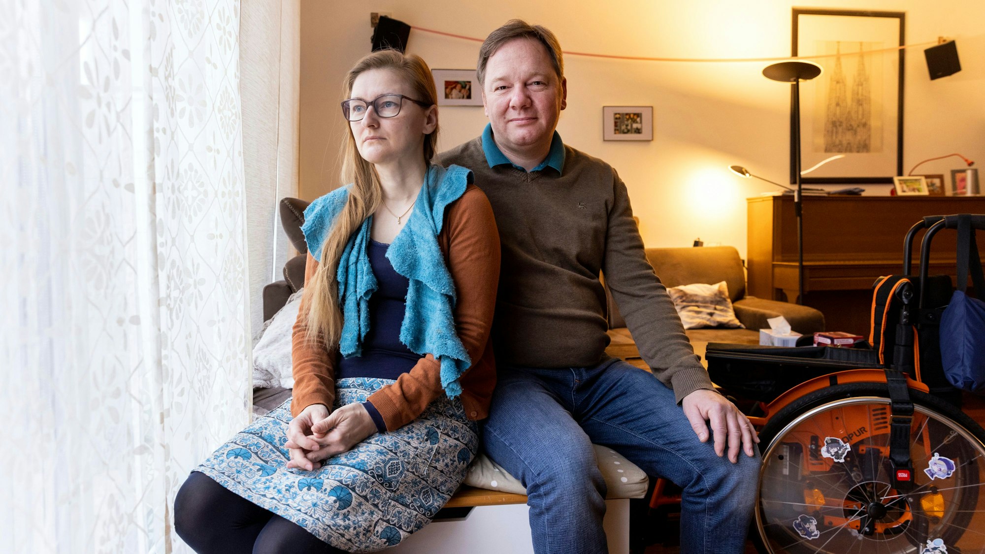 Tanja Bock-Schweizer und Sebastian Bock sitzen auf der Sofakante in ihrem Wohnzimmer. Neben ihnen steht ein Rollstuhl.