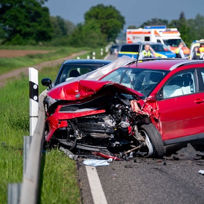 Ein zerstörter roter Kleinwagen steht auf einer Straße bei Euskirchen. Die Fahrerin ist bei dem Unfall ums Leben gekommen. Im Hintergrund sind Polizeifahrzeuge und ein Rettungswagen zu sehen.