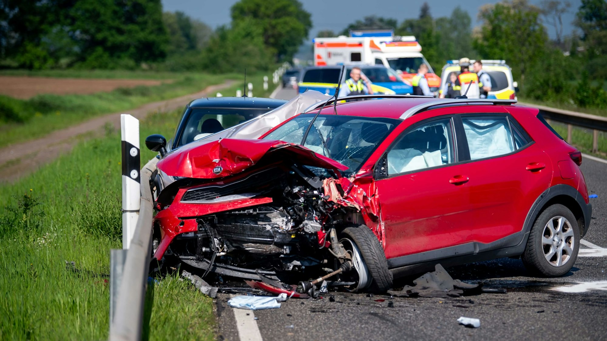 Ein zerstörter roter Kleinwagen steht auf einer Straße bei Euskirchen. Die Fahrerin ist bei dem Unfall ums Leben gekommen. Im Hintergrund sind Polizeifahrzeuge und ein Rettungswagen zu sehen.