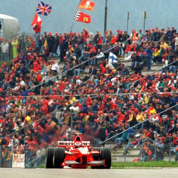 Der rote Rennwagen von Michael Schumacher fährt 1998 vor einer Tribüne voller Fans mit roten Käppis.
