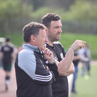 Zwei Trainer stehen am Rand eines Fußballfeldes nebeneinander.