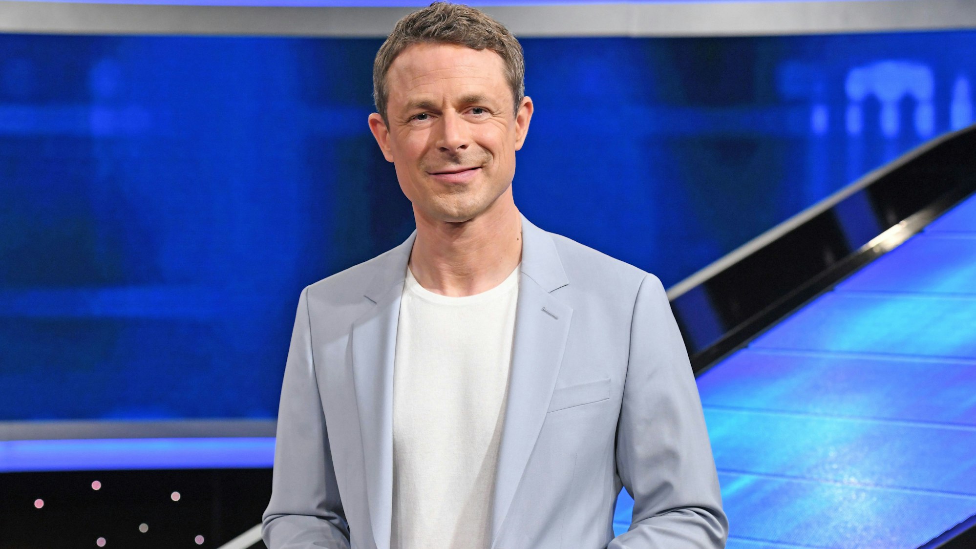 Moderator Alexander Bommes steht vor einer blauen Wand im Studio der ARD-Quizshow „Gefragt – Gejagt“.