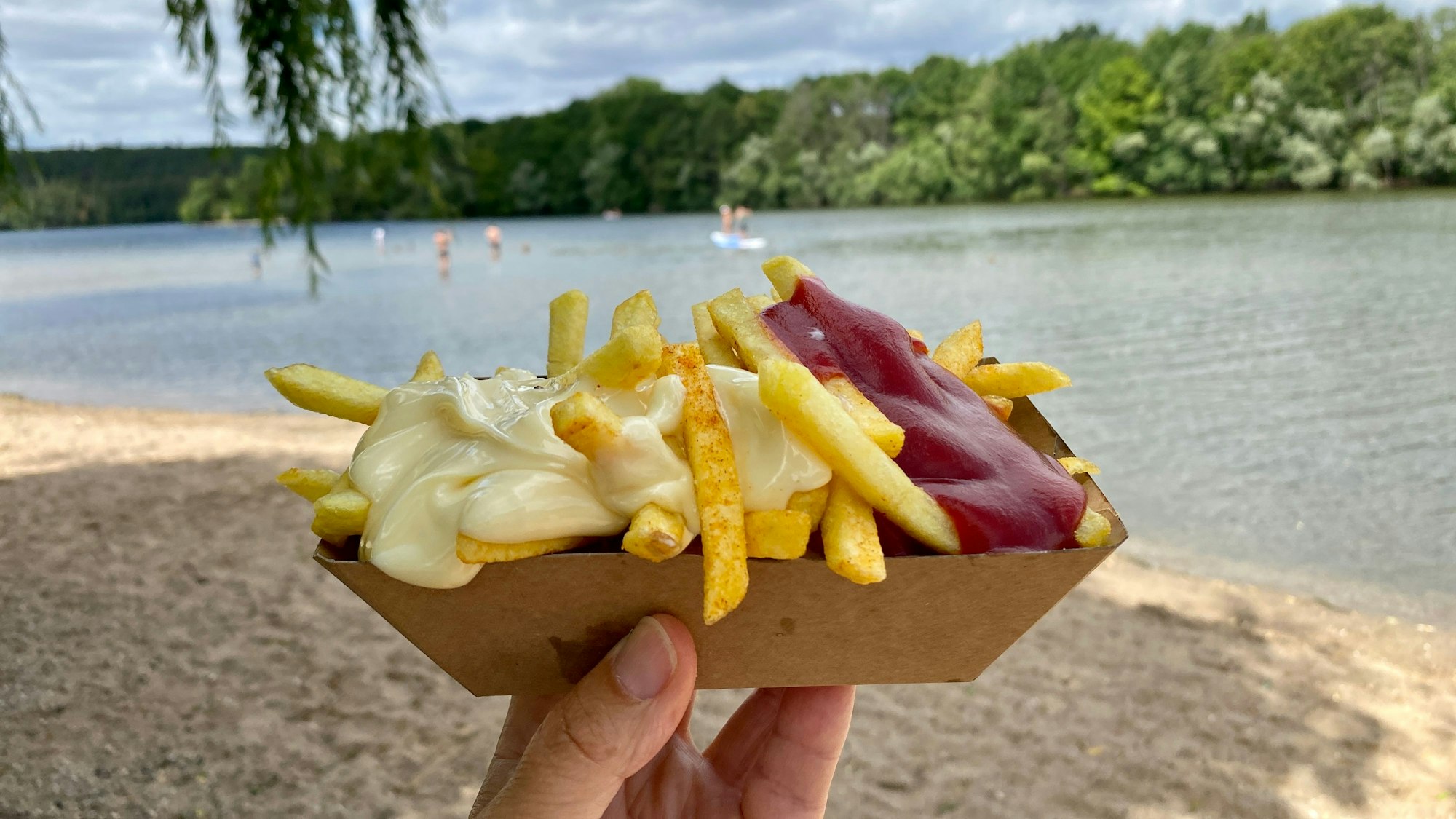 Eine Person hält eine Portion Pommes mit Ketchup und Mayo in der Hand. Im Hintergrund sieht man den Heider Bergsee, in dem Menschen schwimmen und spielen.