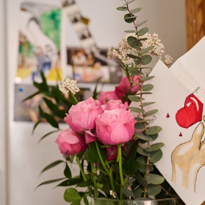 Eine selbst gemalte Karte mit Herz steckt in einem Blumenstrauß, während auf einem Kühlschrank Fotos einer Familie hängen.
Dieser Blumenstrauß und die Karte sind zum Muttertag.