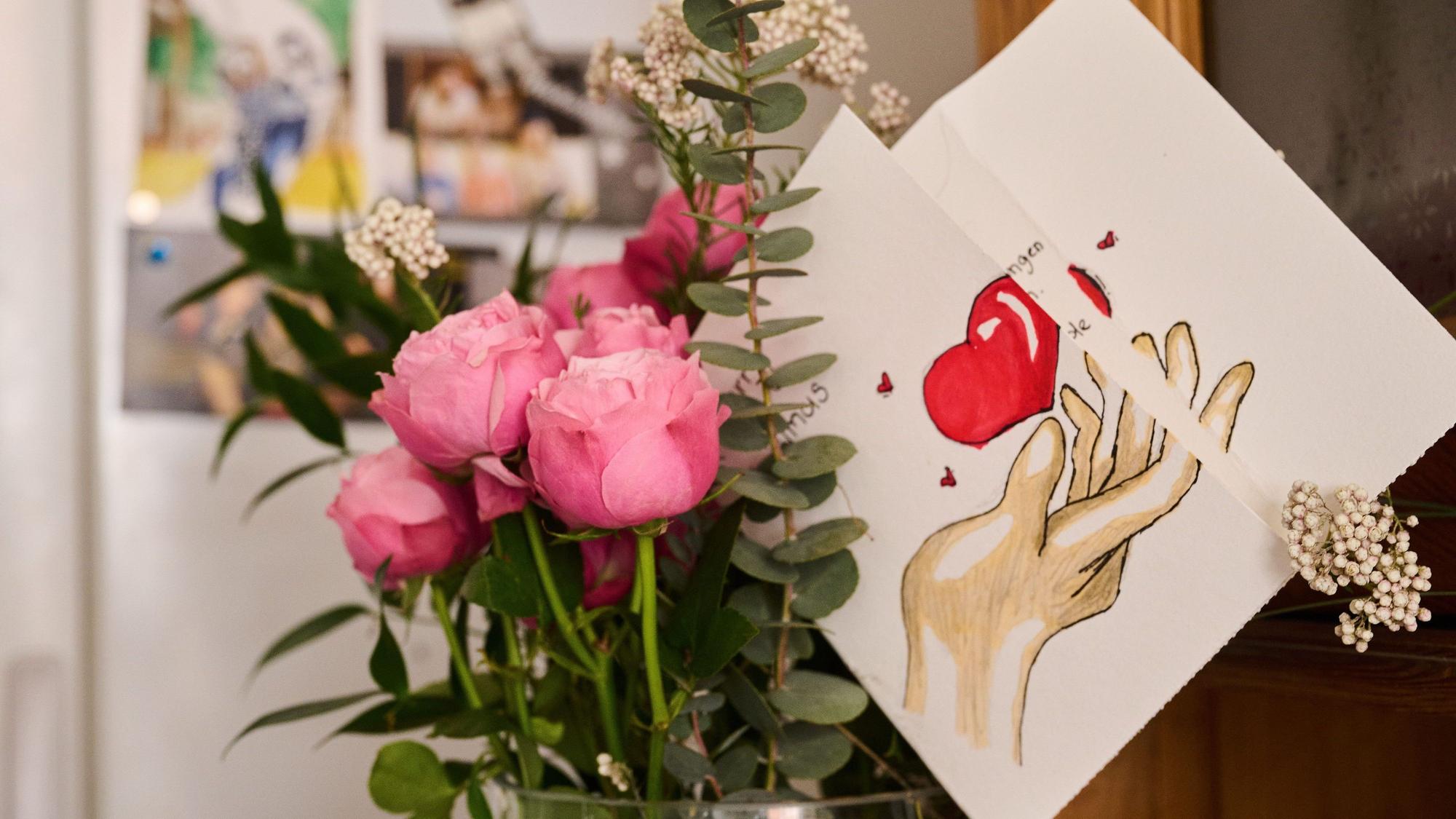 Eine selbst gemalte Karte mit Herz steckt in einem Blumenstrauß, während auf einem Kühlschrank Fotos einer Familie hängen.
Dieser Blumenstrauß und die Karte sind zum Muttertag.