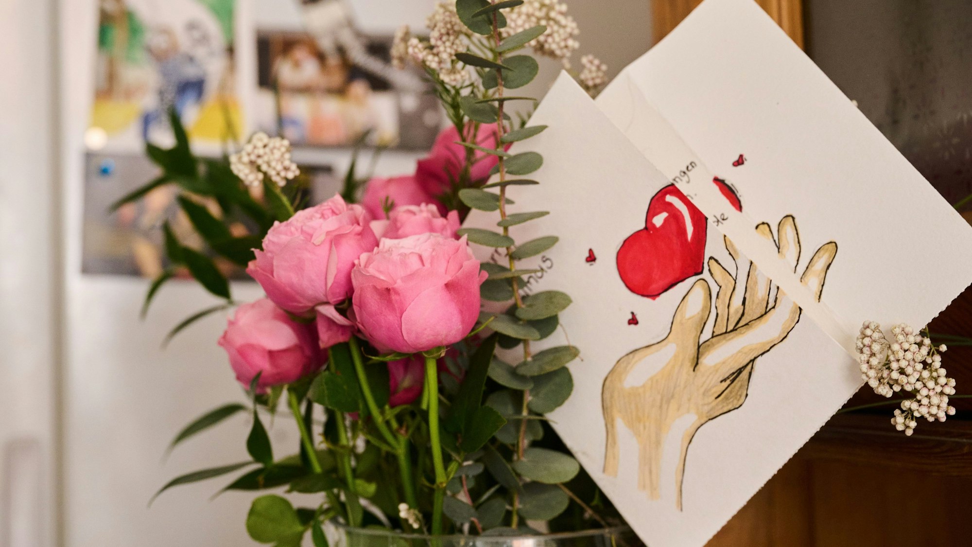 Eine selbst gemalte Karte mit Herz steckt in einem Blumenstrauß, während auf einem Kühlschrank Fotos einer Familie hängen.
Dieser Blumenstrauß und die Karte sind zum Muttertag.