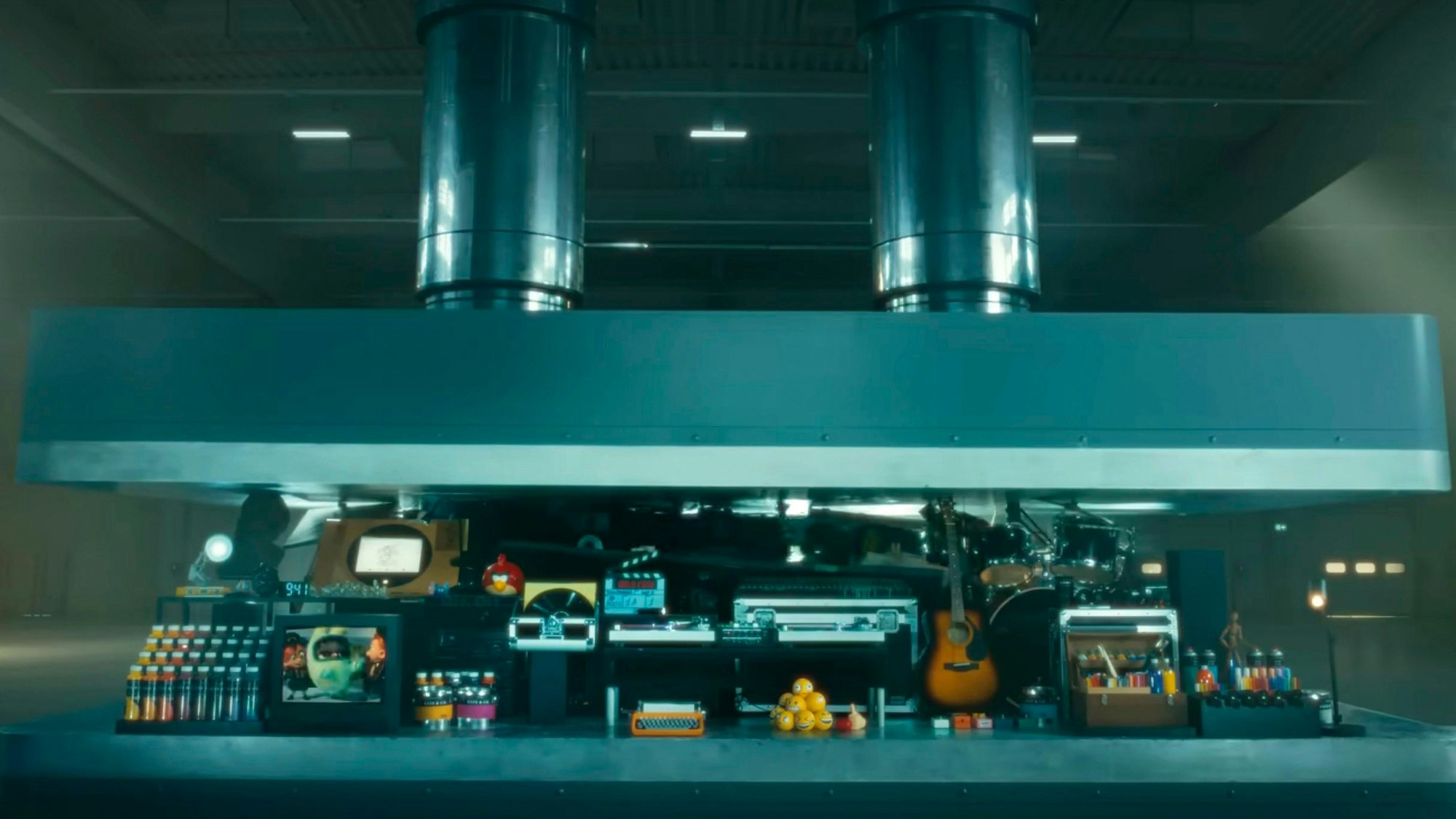 In der oberen Bildhälfte ist eine große graue Presse zu sehen, die sich langsam gen Boden senkt. Dazwischen reihen sich unzählige Gegenstände, zum Beispiel Farbeimer, Musikinstrumente und Emoji-Puppen.