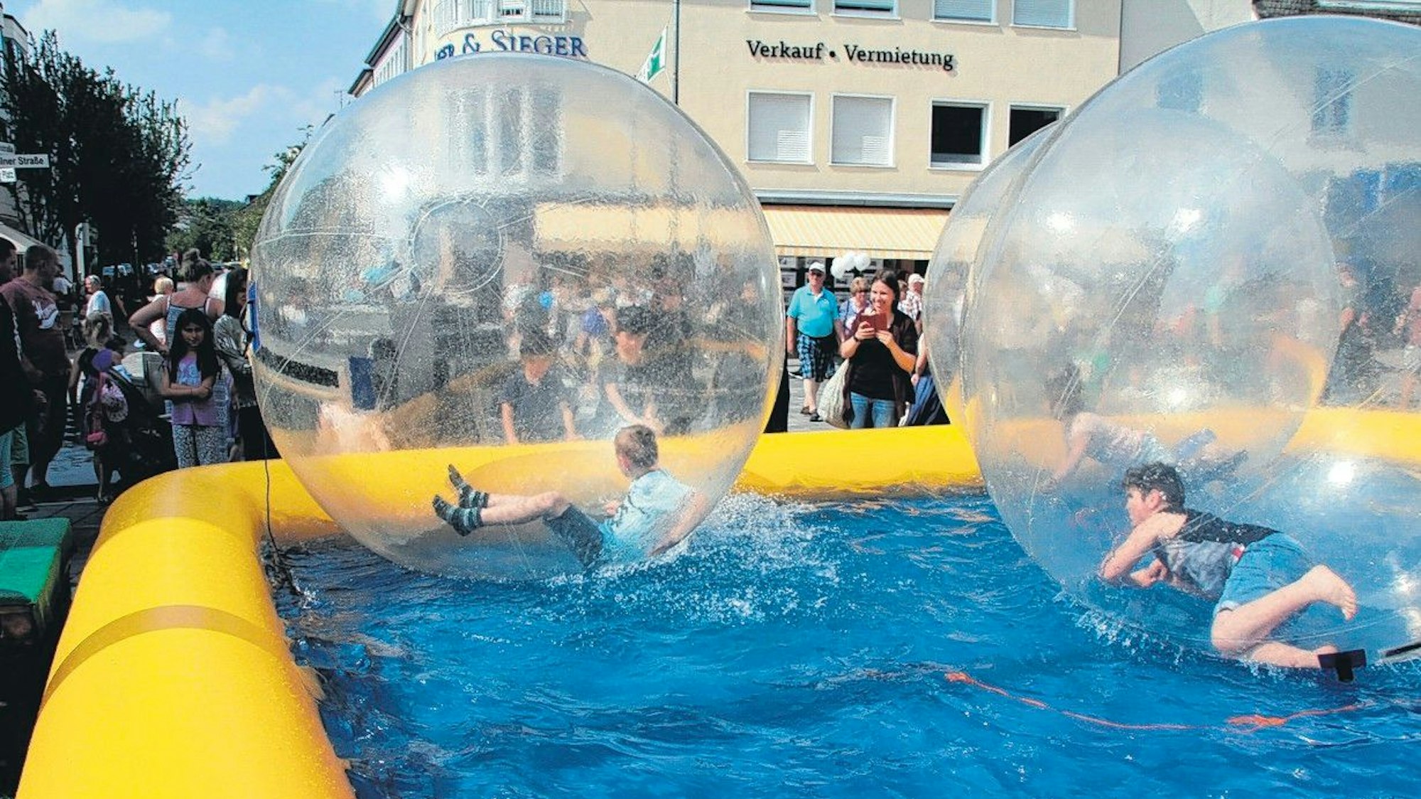 Ein großes Planschbecken in einer Fußgängerzone. In transparenten Kugeln bewegen sich Kinder im Wasser.