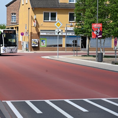 Auf dem Foto ist die Carl-Schurz-Straße in Erftstadt-Liblar zu sehen. Ein Bus fährt um die Ecke. Die gesamte Kreuzung ist mit einem roten Belag versehen.